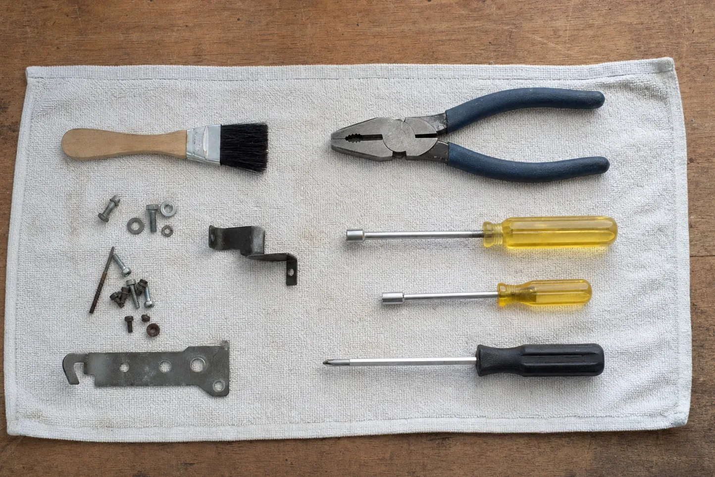 Hand tools, brackets and loose fasteners laid out in rows on a towel over a scratched wooden bench